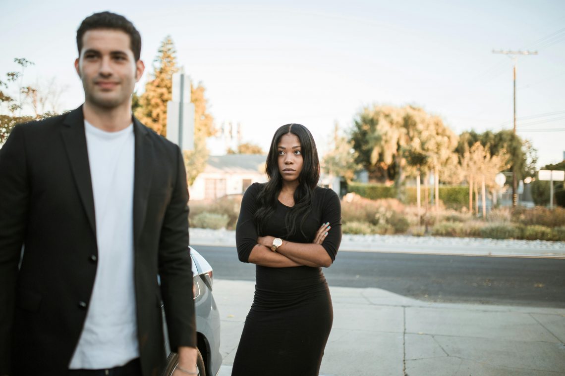 man smirks while his upset girlfriend stands behind him with her arms crossed