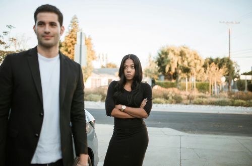 man smirks while his upset girlfriend stands behind him with her arms crossed