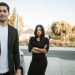 man smirks while his upset girlfriend stands behind him with her arms crossed
